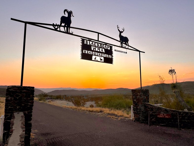A sign hangs over a gate with a sunset in the background