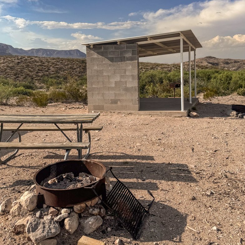 A campsite with a fire ring, picnic table and concrete shade shelter