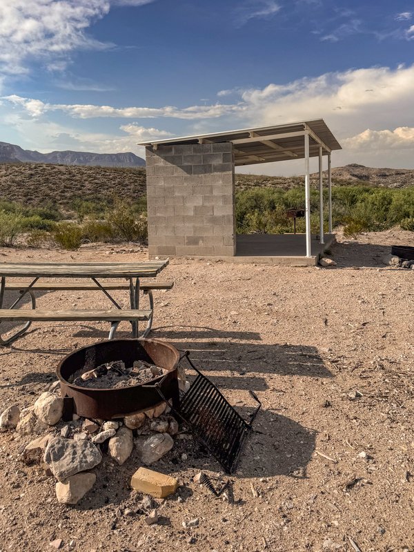 A campsite with a fire ring, picnic table and concrete shade shelter