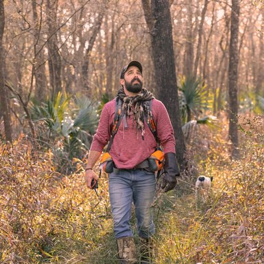 A man in hunting gear walks through a wooded, grassy area with a dog in the background