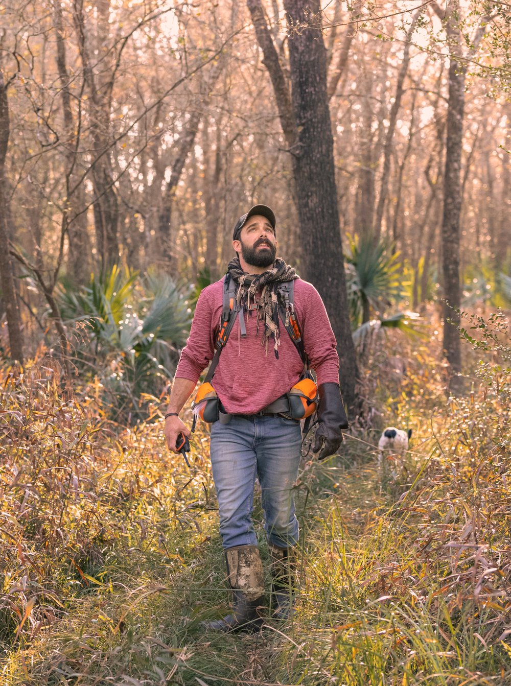 A man in hunting gear walks through a wooded, grassy area with a dog in the background