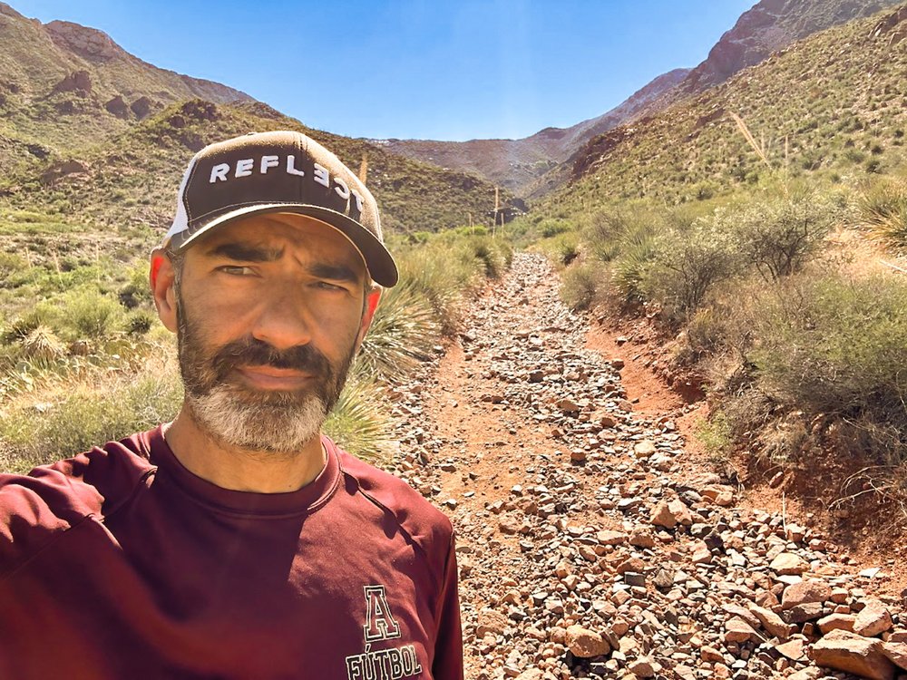 A man wearing a hat takes a selfing along a rocky trail in the mountains.