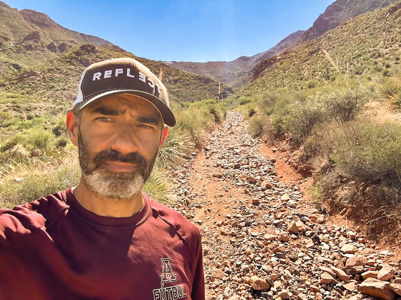 A man wearing a hat takes a selfing along a rocky trail in the mountains.