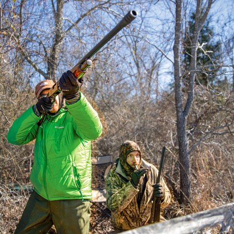 A man in a bright green jacket aims his shotgun at a duck while another man in all camo blows into a duck whistle