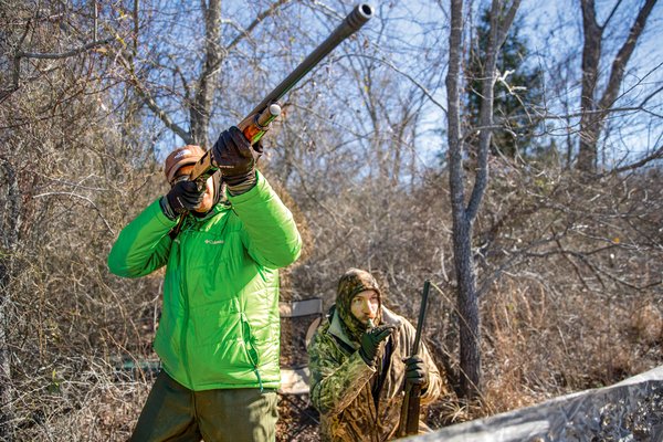 A man in a bright green jacket aims his shotgun at a duck while another man in all camo blows into a duck whistle