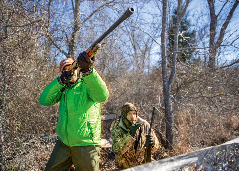 A man in a bright green jacket aims his shotgun at a duck while another man in all camo blows into a duck whistle