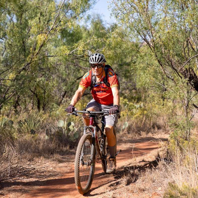 A man wearing a red shirt bikes along a trail