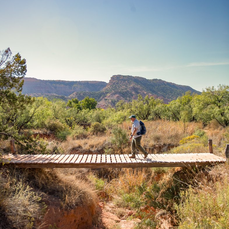 A man with treking poles walks across a wooden bridge over a dry creek bed at Palo Duro Canyon State Park