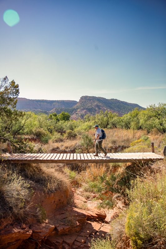 A man with treking poles walks across a wooden bridge over a dry creek bed at Palo Duro Canyon State Park