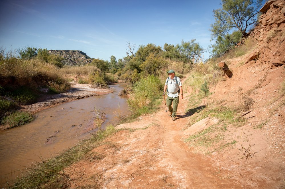 A man with a treking pole hikes along a creek on the Paseo del Rio Trail at Palo Duro Canyon State Park