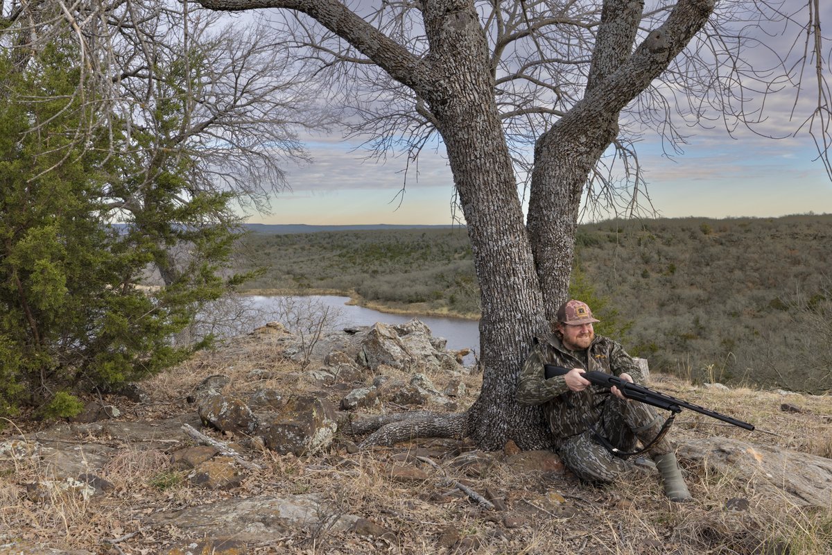 Hunter pointing a gun while resting against a tree.
