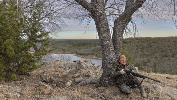 Hunter pointing a gun while resting against a tree.