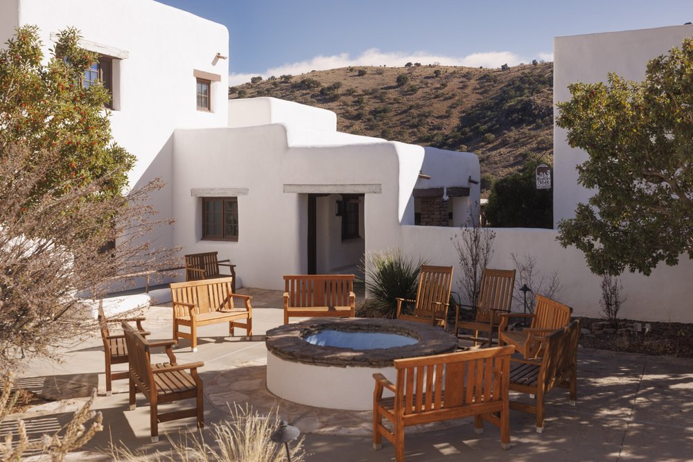 A white adobe lodge, featuring a courtyard with wooden chair seating around a firepit