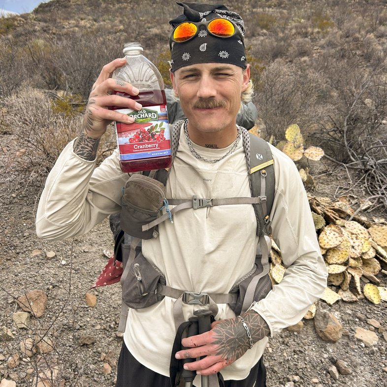 A man in hiking gear holding a bottle of cranberry juice on the trail