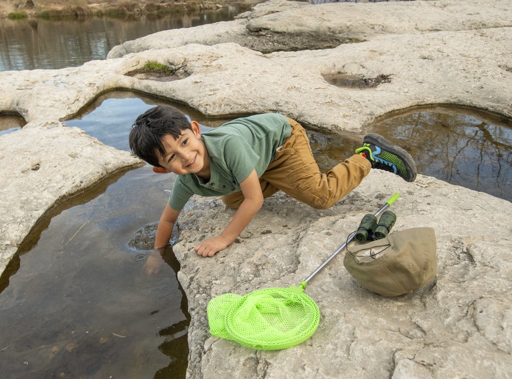 A child with a net and other supplies reaching his hand in the water between rock formations