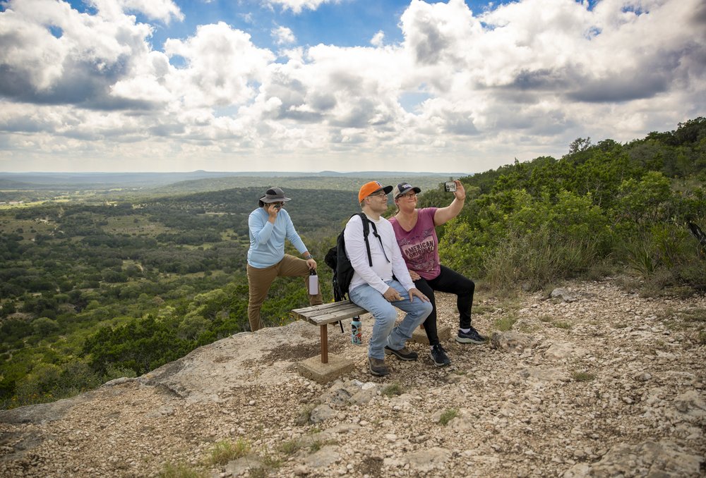 Two people sitting on a bench with a person behind them taking a selfie in the hill country.