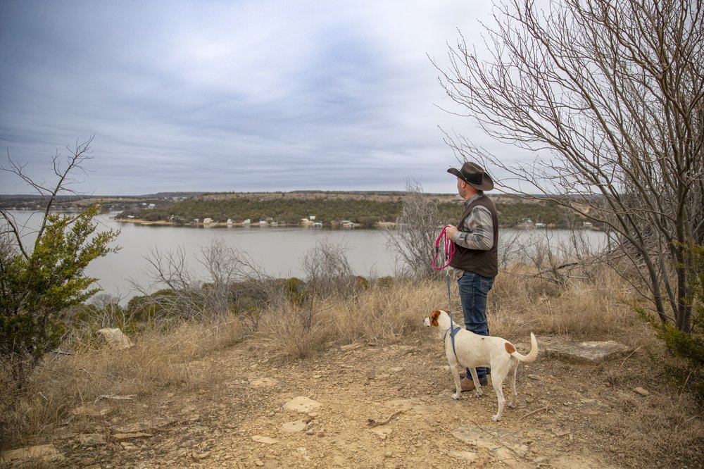 A man in a brown cowboy hat poses at a scenic overlook with his white and brown dog