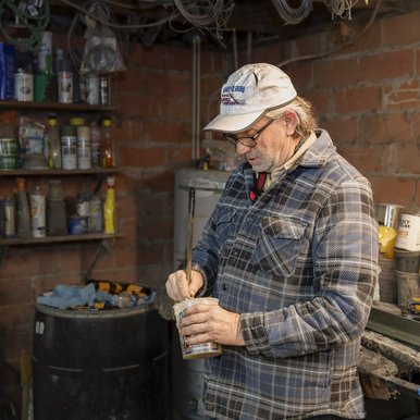 Canoe builder stirring epoxy in a can in front of his workbench lined with canoe building equipment