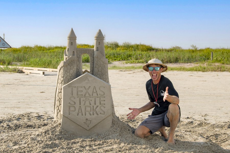 A man with an excited smile poses next to a sand castle that has the Texas State Parks logo carved into it
