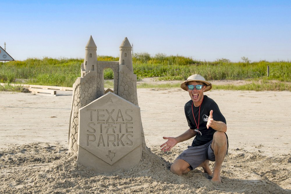 A man with an excited smile poses next to a sand castle that has the Texas State Parks logo carved into it