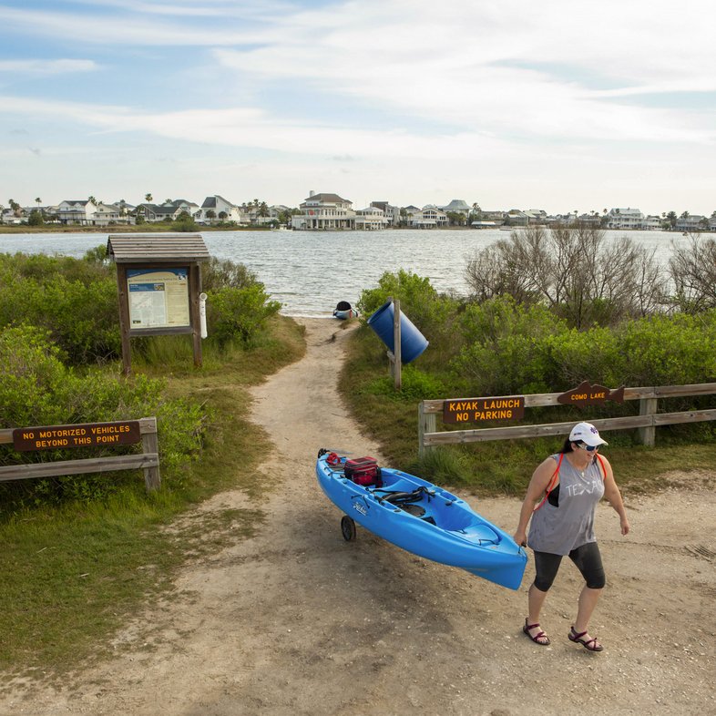 Female paddler is rolling a blue kayak away from launching area off the Galveston Bay