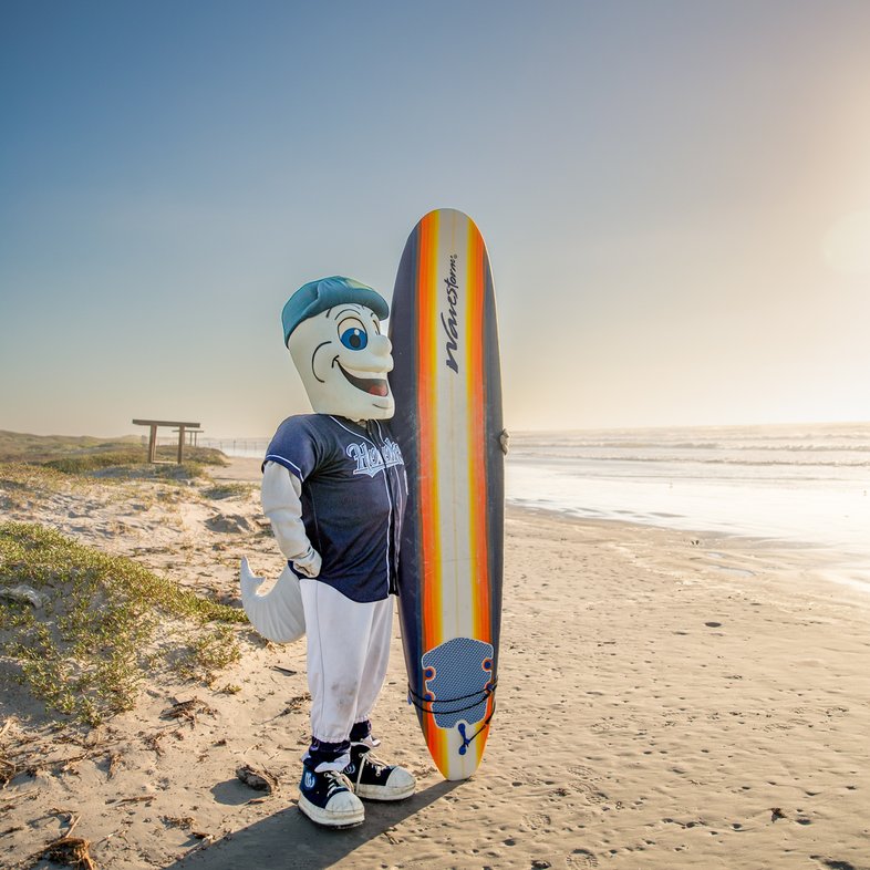 Corpus Christi Hooks mascot Rusty on the beach at Mustang Island State Park holding a surfboard.