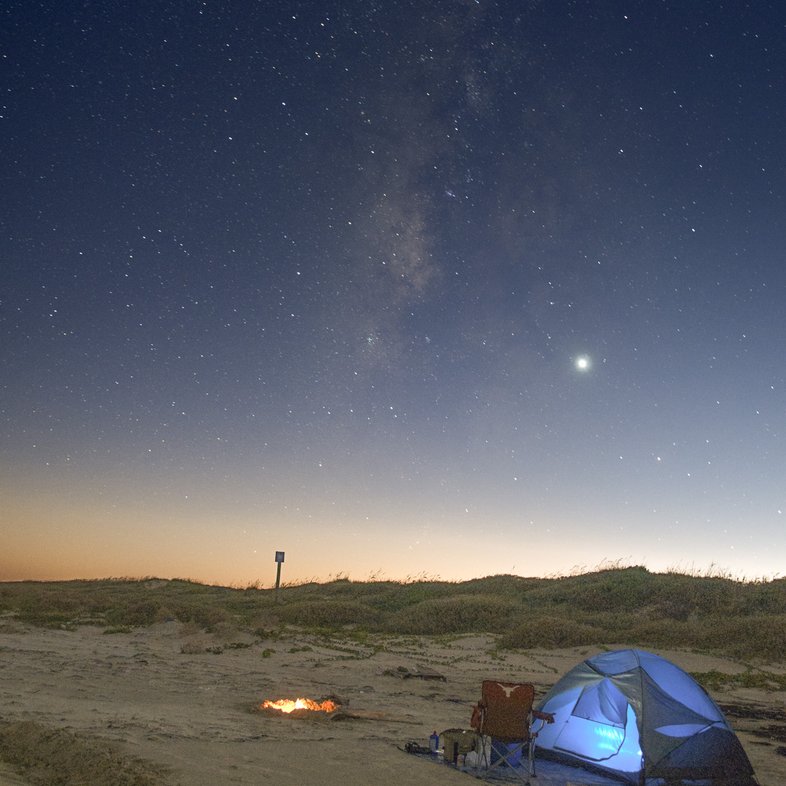 An illuminated blue dome tent pitched on the beach next to a campfire at dusk on Padre Island National Seashore