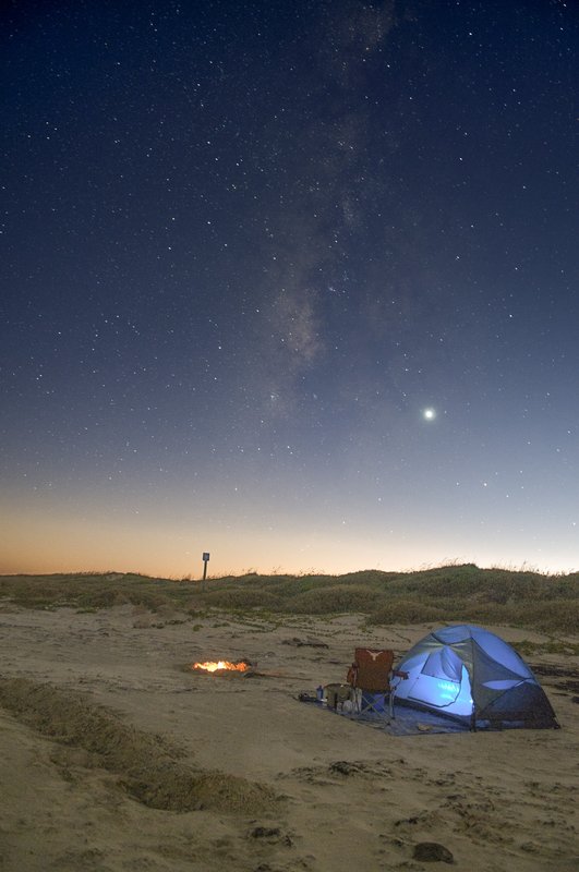 An illuminated blue dome tent pitched on the beach next to a campfire at dusk on Padre Island National Seashore