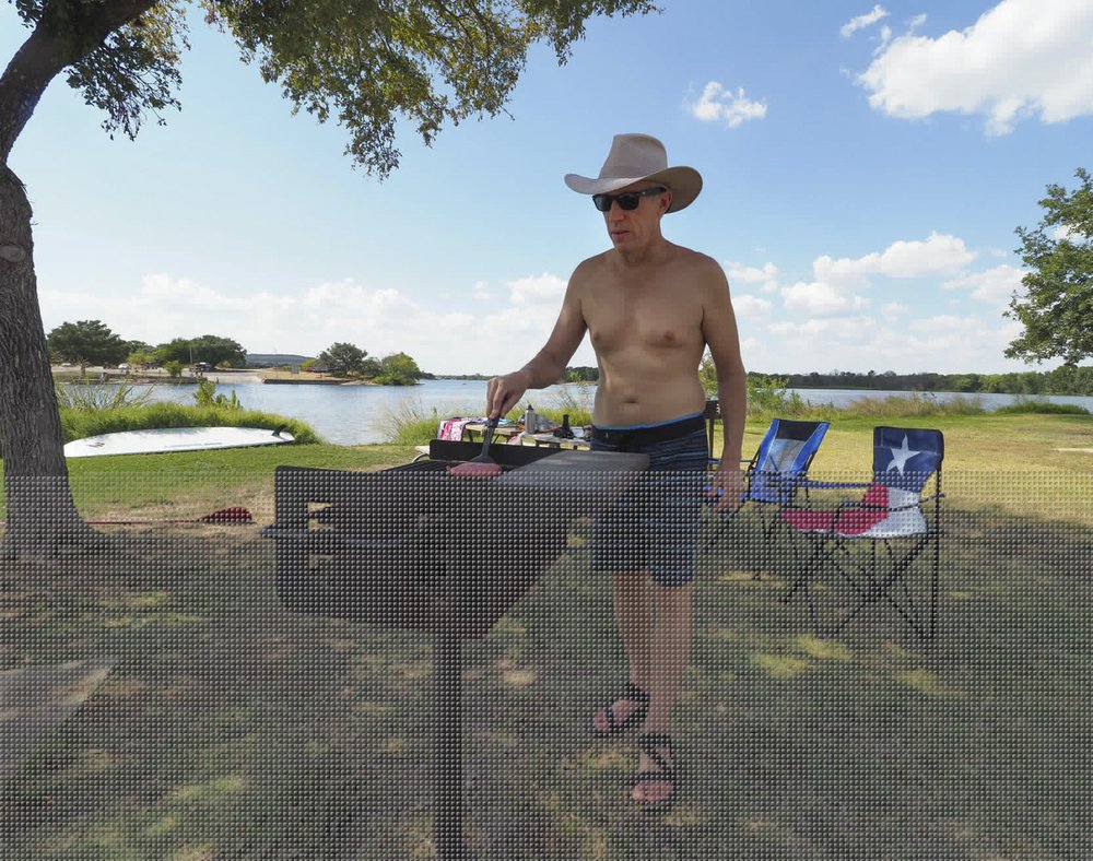 A shirtless man in a cowboy hat grills lunch over a charcoal stove
