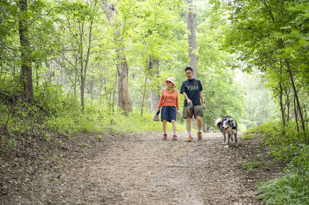Two hikers in shorts walk their dog along the tree-lined Bald Cypress trail at Guadalupe River State Park