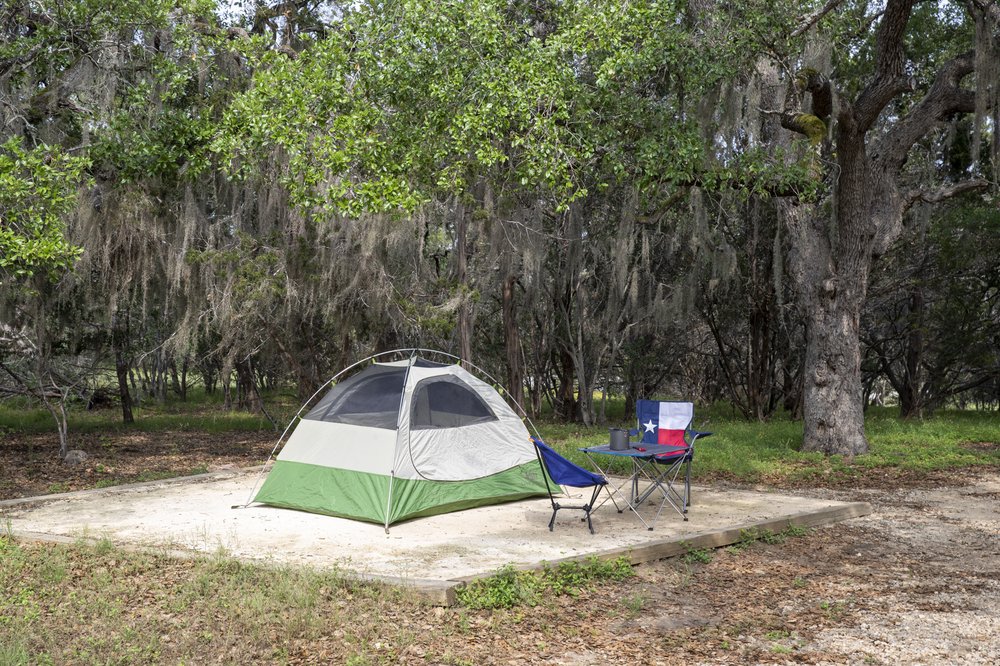 A green and tan dome tent pitched on a square dirt camping area surrounded by grass