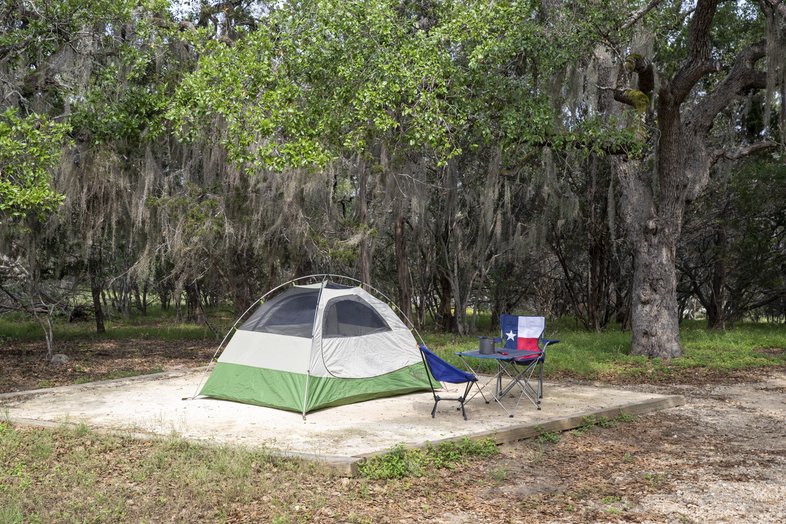 A green and tan dome tent pitched on a square dirt camping area surrounded by grass