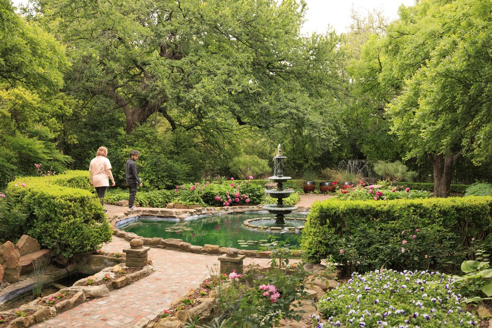 Two people walk along a paved garden path past a fountain in Weatherford's Chandor Gardens