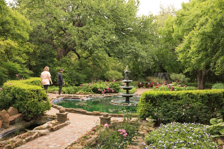 Two people walk along a paved garden path past a fountain in Weatherford's Chandor Gardens