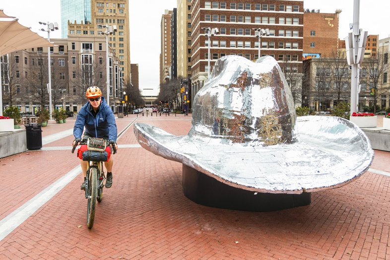 A person riding a bike by a silver cowboy hat sculpture.
