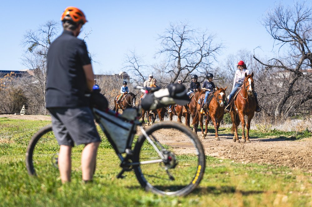 A person with a bike standing near a trail with people on horseback.