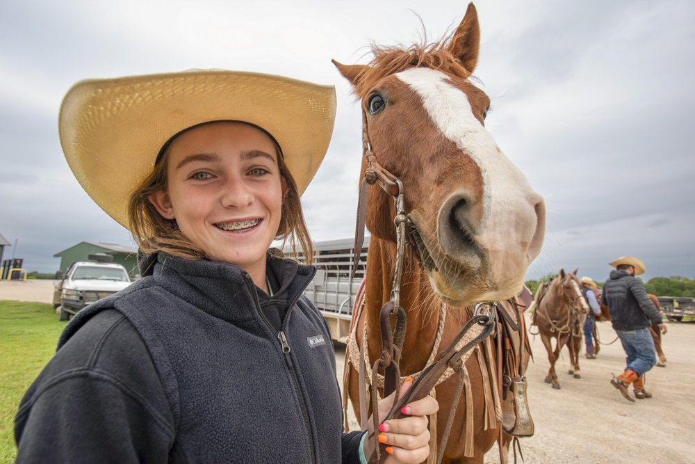A young woman wearing a cowboy hat holds the reins of her saddled horse.