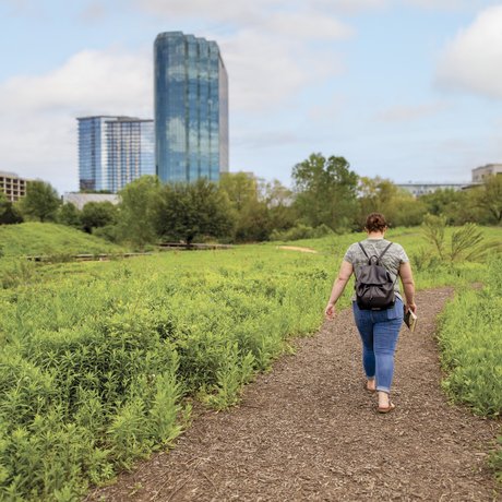 A woman walks a bark-covered path in an urban greenbelt, with tall buildings in the background