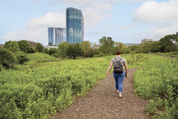 A woman walks a bark-covered path in an urban greenbelt, with tall buildings in the background