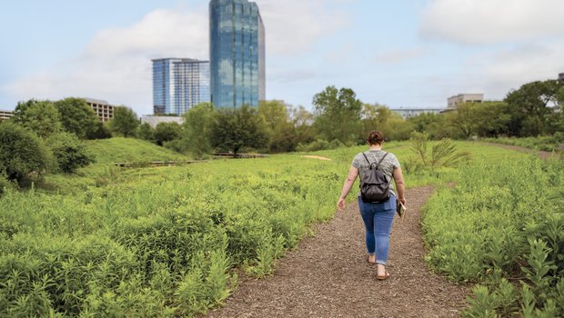 A woman walks a bark-covered path in an urban greenbelt, with tall buildings in the background