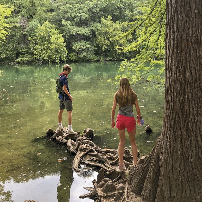Two kids walk along the gnarled roots of a tree out over the water of a river
