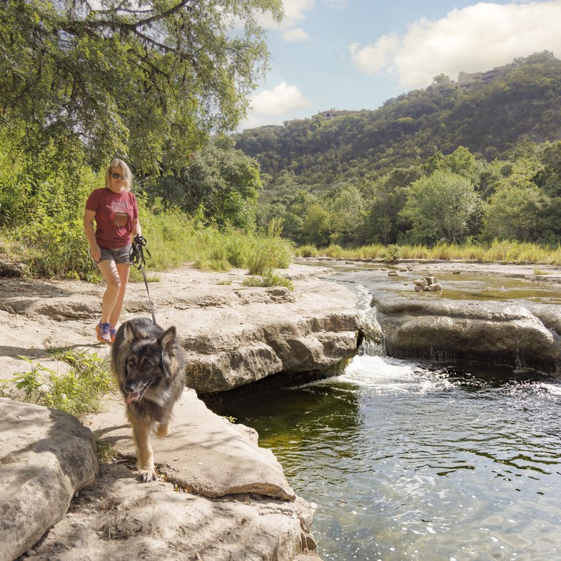 A woman walks her dog along the rocky shore of a river with a small waterfall in the background