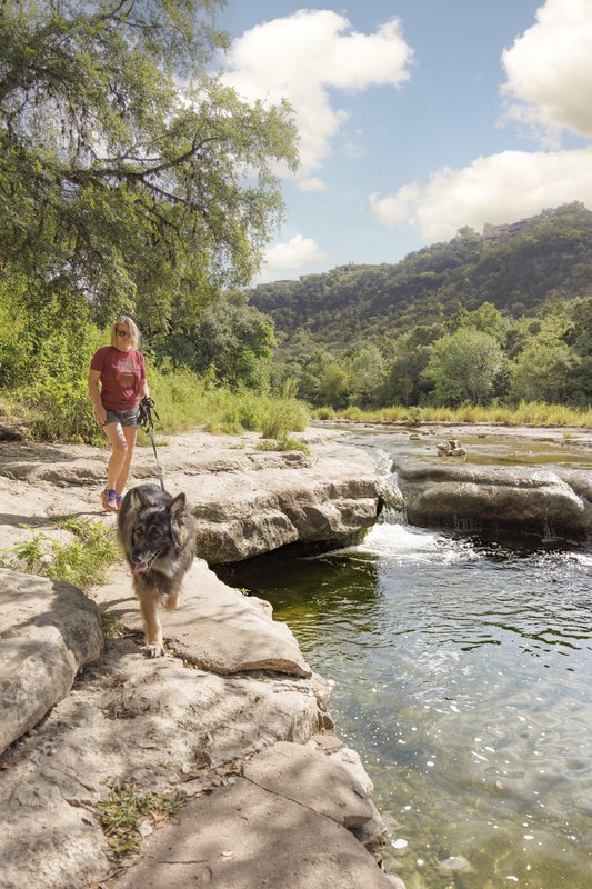 A woman walks her dog along the rocky shore of a river with a small waterfall in the background