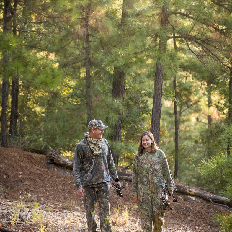 A man and woman both dressed in camouflage and holding compound bows walk along a trail