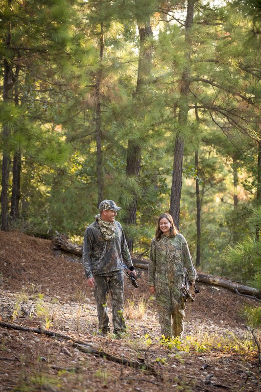 A man and woman both dressed in camouflage and holding compound bows walk along a trail