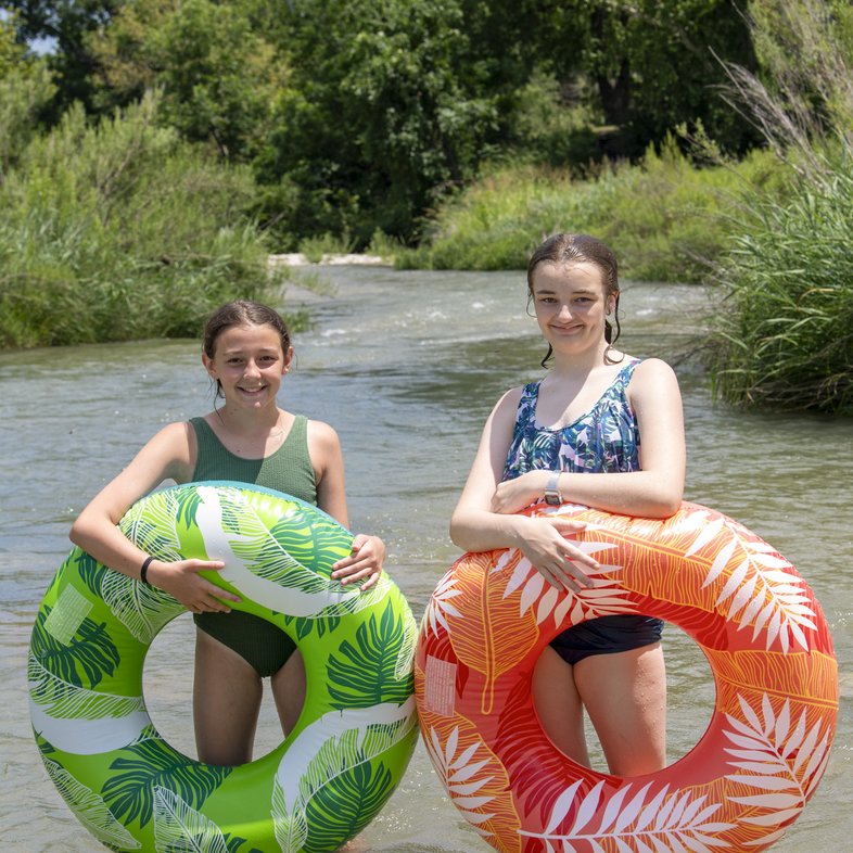 Two young women holding inflatable tubes stand in the water of the South Llano River in Junction Texas