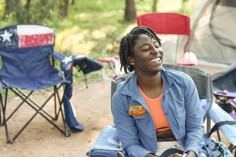 An African American woman smiles while sitting on a camp chair in front of a tent