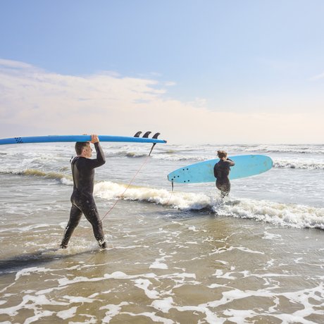 A father and daughter, both dressed in black wetsuits, carry blue surfboards into the water at Mustang Island