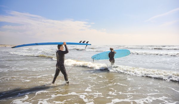 A father and daughter, both dressed in black wetsuits, carry blue surfboards into the water at Mustang Island