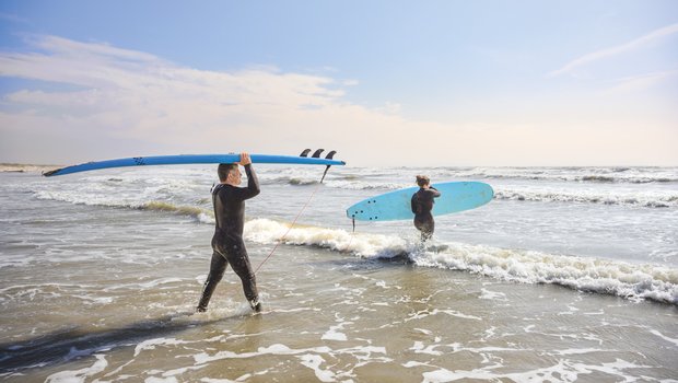 A father and daughter, both dressed in black wetsuits, carry blue surfboards into the water at Mustang Island